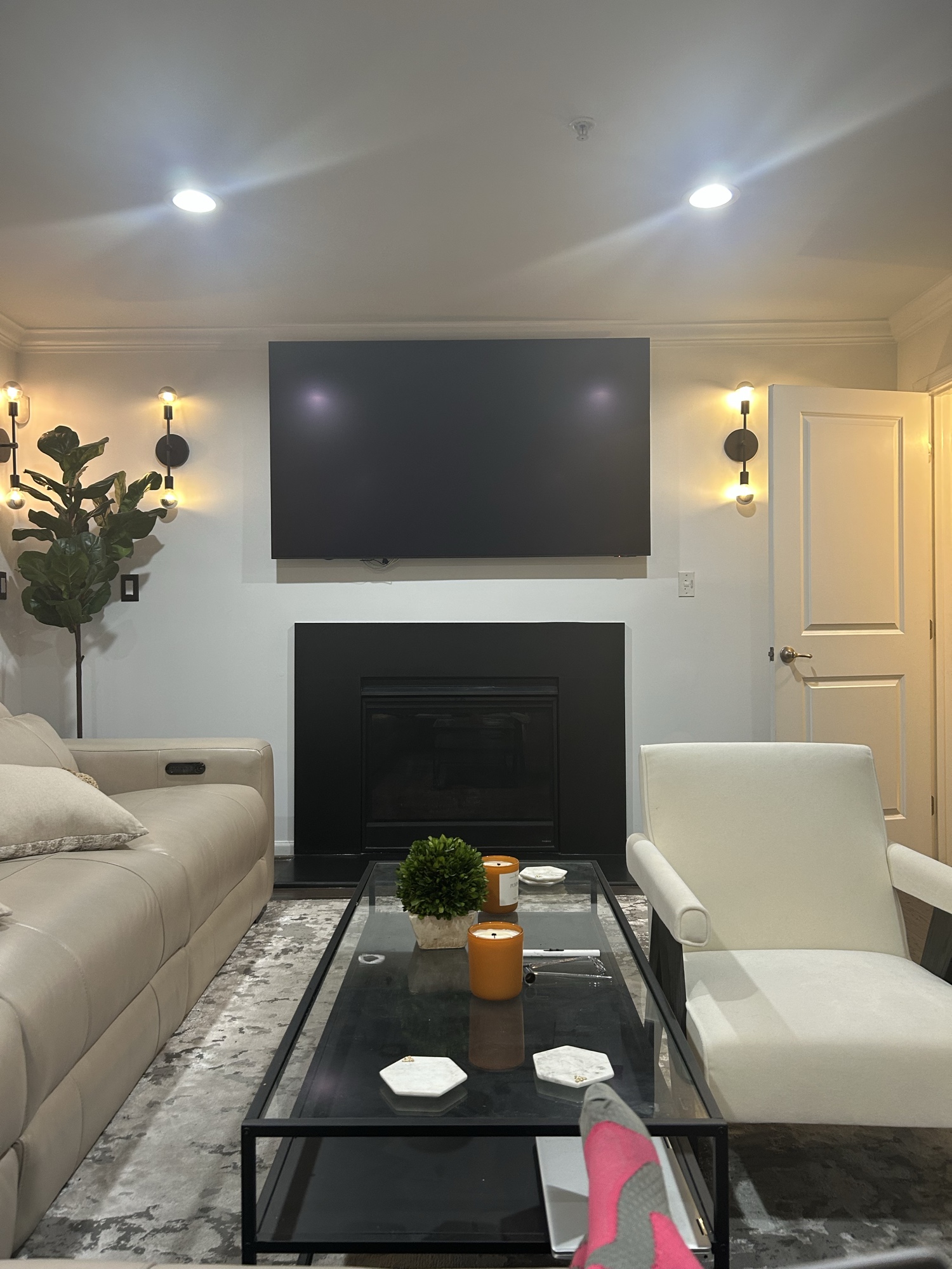 After: styled living room with curved cream sectional, glass coffee table, twin wall sconces, candles, and a fiddle leaf fig