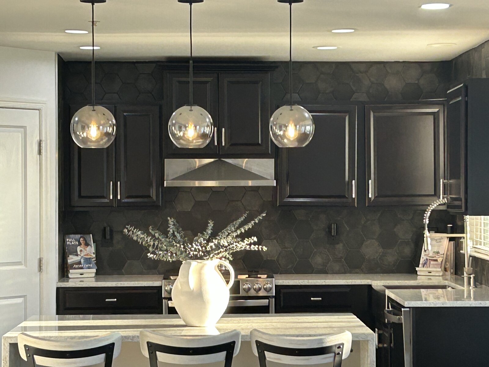 Detail: matte-black hexagon backsplash, stainless range hood, smoke-glass globe pendants centered on the black cabinetry