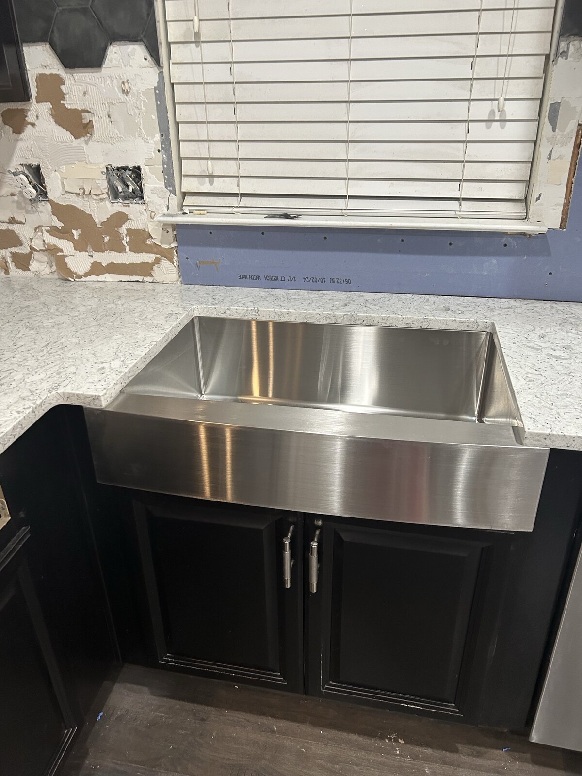 During install: new stainless apron-front farmhouse sink set into fresh quartz counter against raw drywall