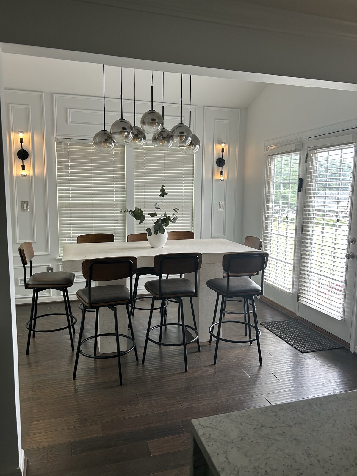 Dining room with double-row globe pendant chandelier, long quartz table, six leather and wood barstools, picture-frame moulding, twin black sconces, and French doors to a sunlit garden