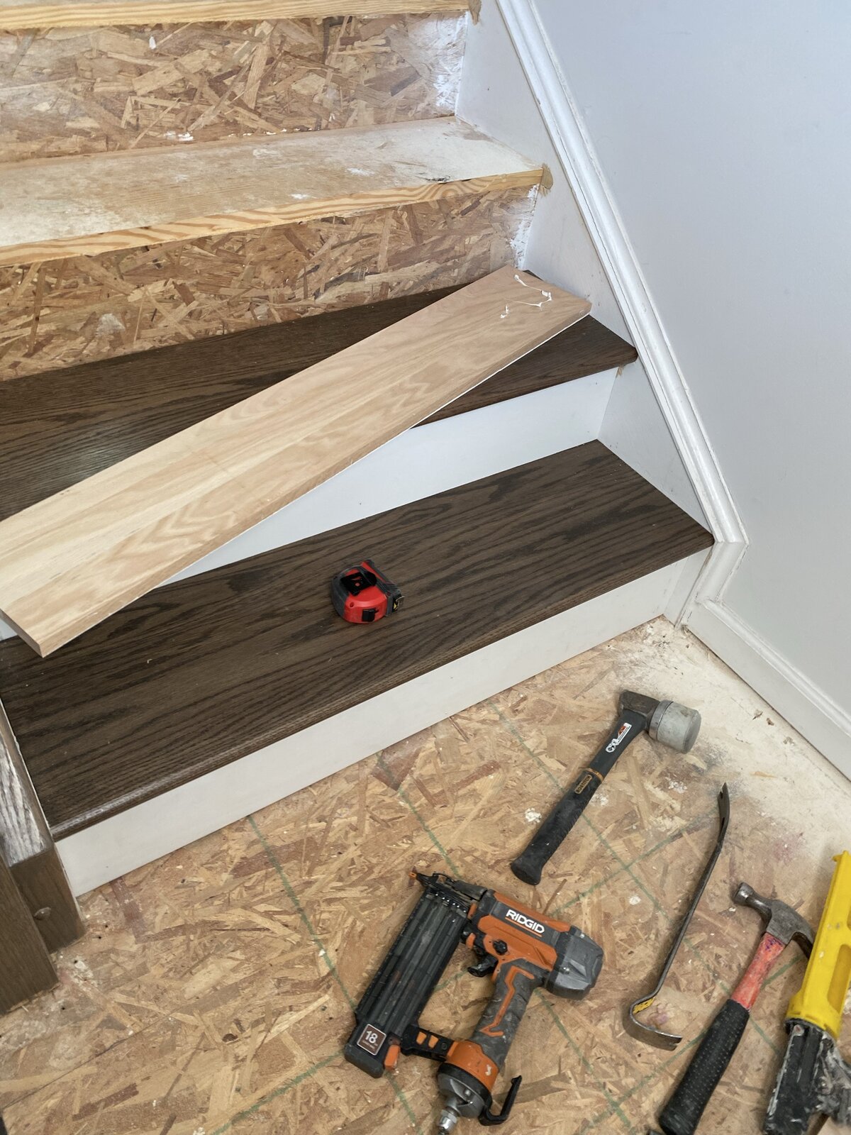Staircase mid-renovation — solid oak tread being nailed in place with a Ridgid pneumatic finish nailer, tools and level visible on the stairs